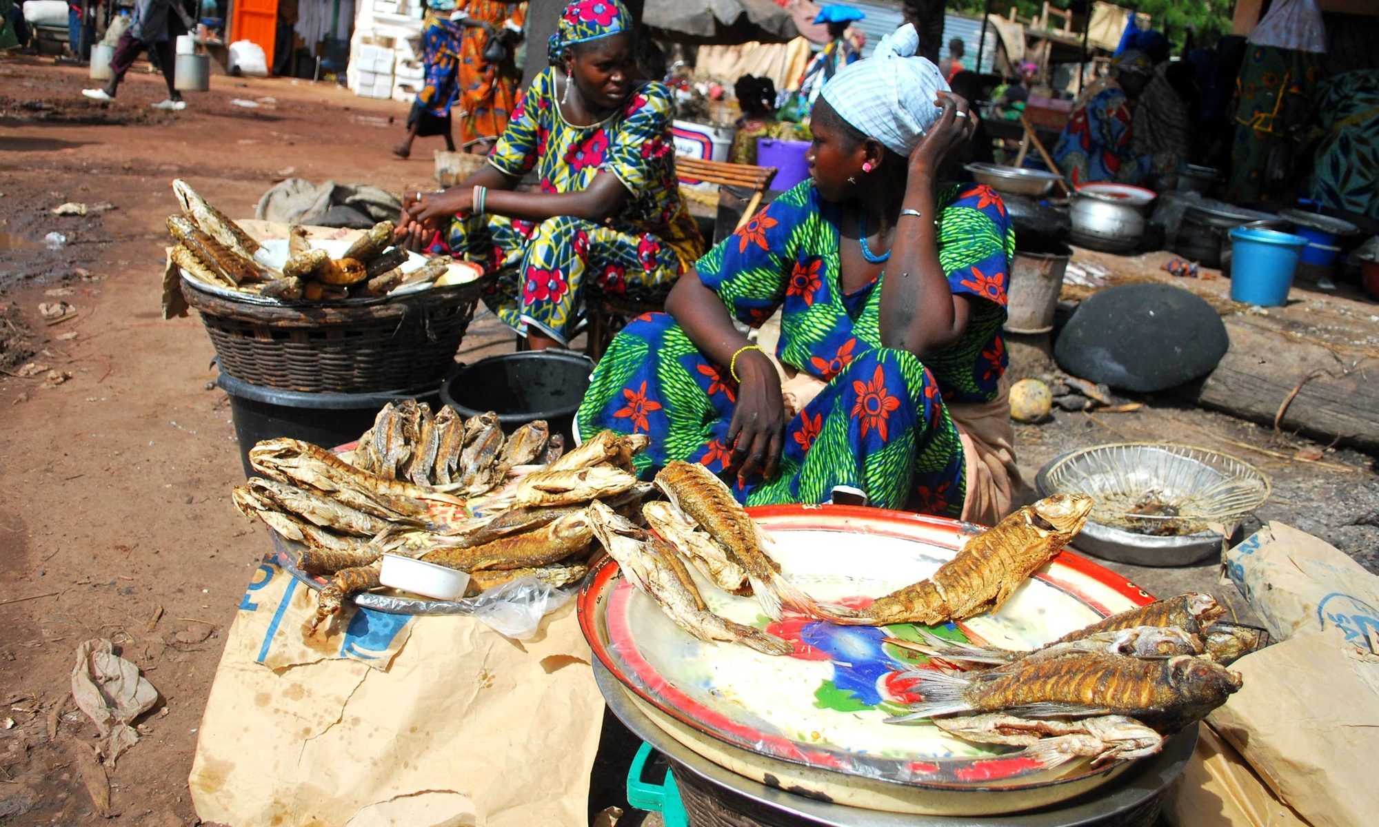 A trader at her stall, smoked fish laid out on an enamel tray in an open-air West African market
