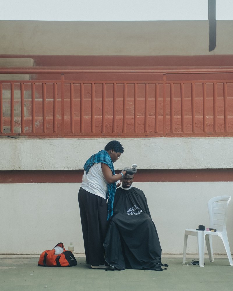 A roadside barber in Ibadan cutting a customer's hair outdoors against a long painted wall