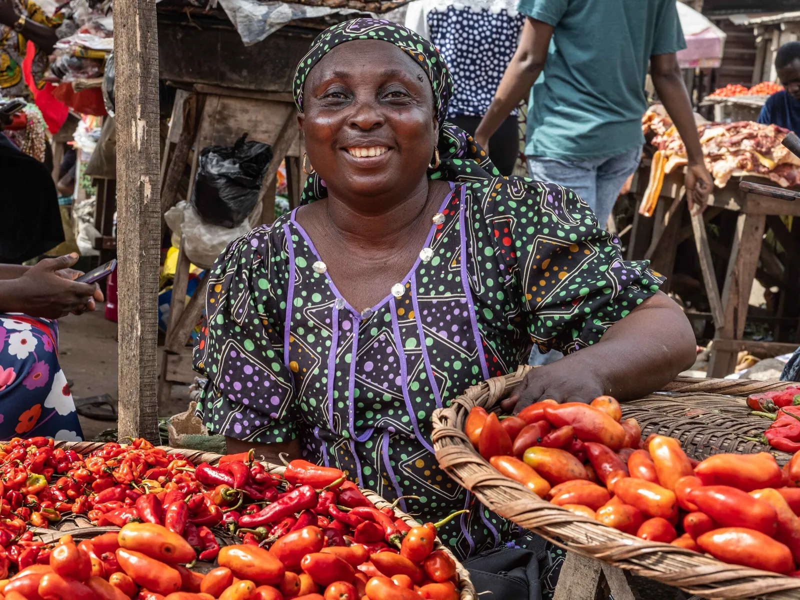 A smiling woman at her market stall behind baskets of red peppers and tomatoes in Ogun State