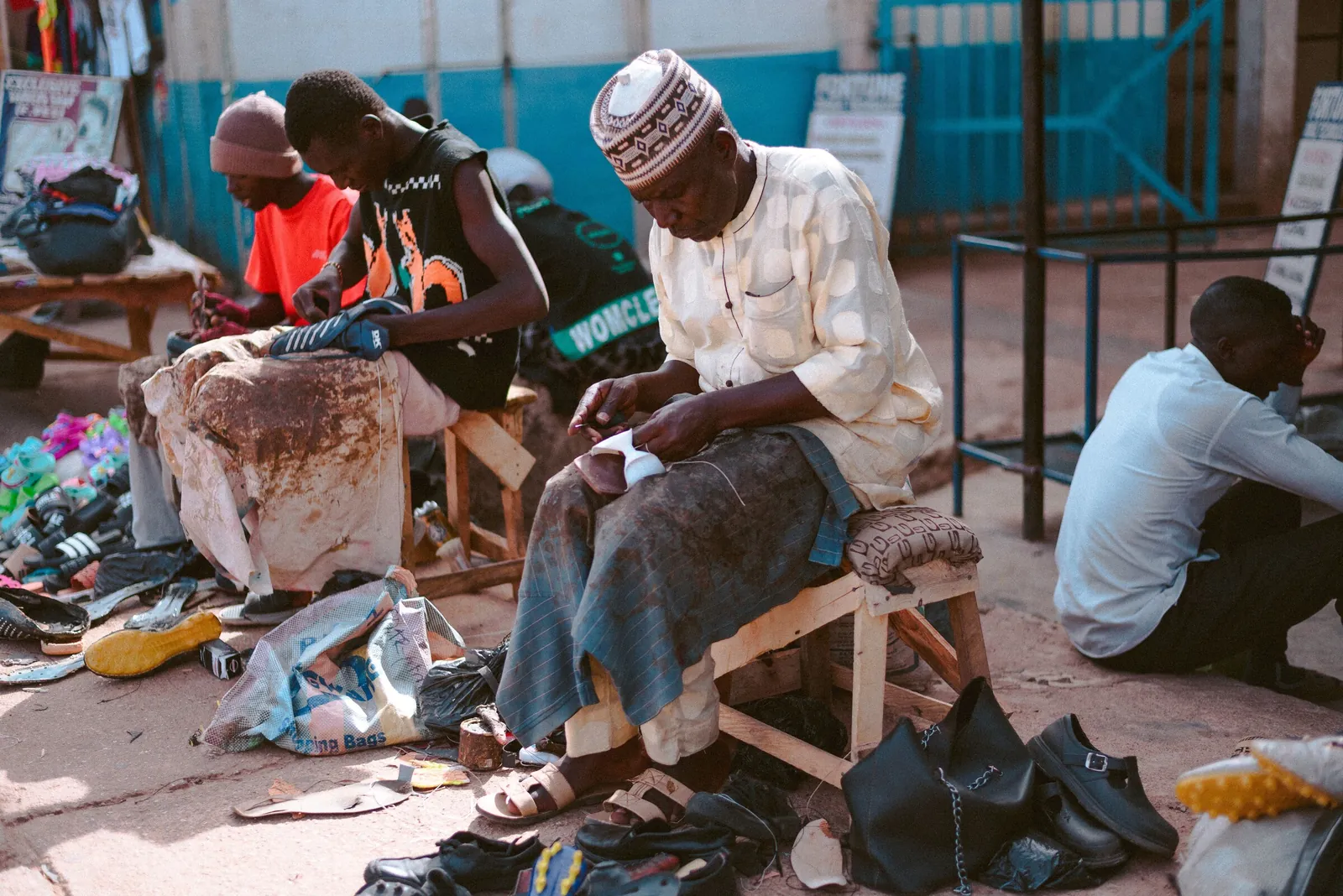 Three shoemakers at work on low stools in an open-air market in Jos